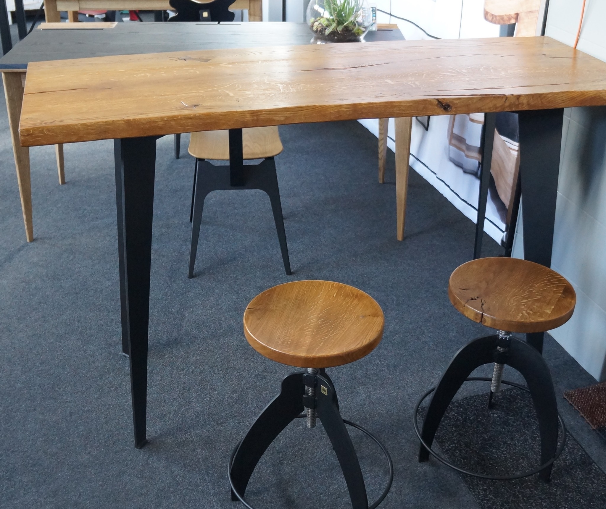 Wysoki stół z blatem z jasnego drewna o widocznej strukturze słojów, wsparty na czarnych, smukłych nogach, oraz dwa drewniane hokery z regulowaną wysokością na metalowej podstawie, na tle szarej...