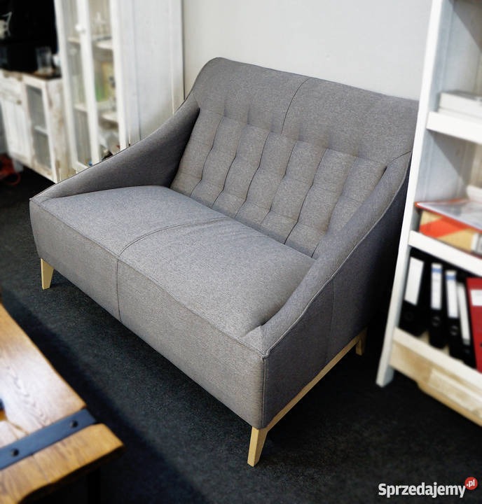 Szara sofa dwuosobowa z tapicerowanym oparciem w pionowe kwadraty, na jasnych drewnianych nóżkach, widoczna w pomieszczeniu z meblami w tle.