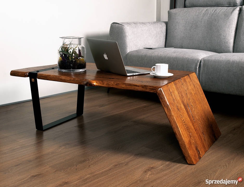 Stolik kawowy z blatem z litego drewna o nieregularnych krawędziach, wsparty na metalowych nogach i drewnianej podstawie, na blacie laptop, filiżanka kawy i szklany wazon z roślinami, w tle szara...