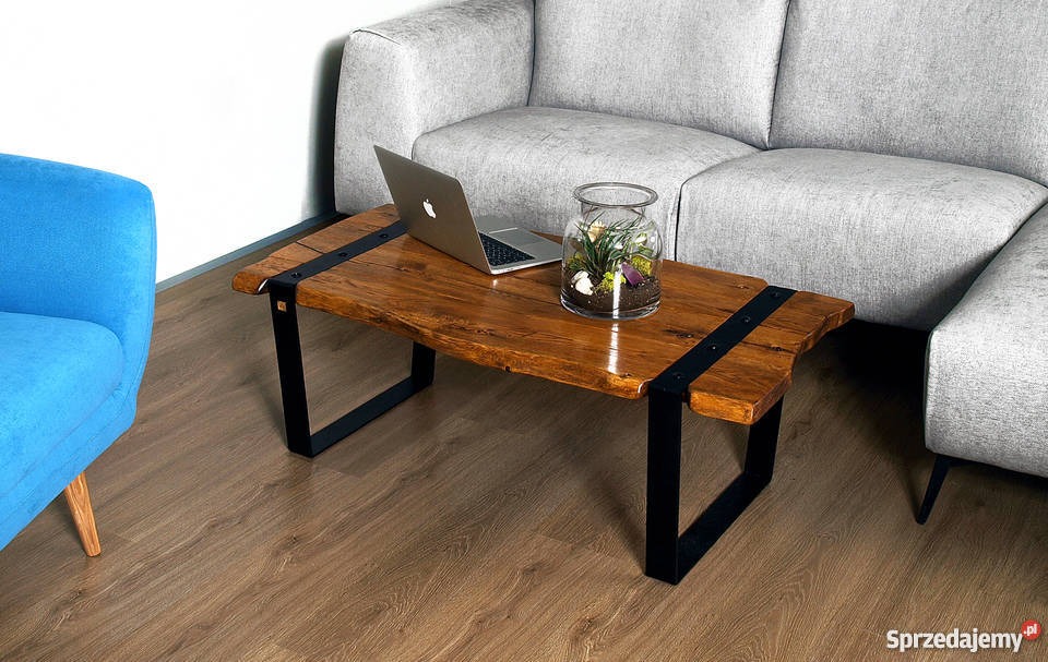 Stolik kawowy z blatem z litego drewna o nieregularnych krawędziach, wzmocniony czarnymi metalowymi paskami i nogami, na blacie laptop i szklany wazon z roślinami, w tle szara sofa i błękitny fotel.