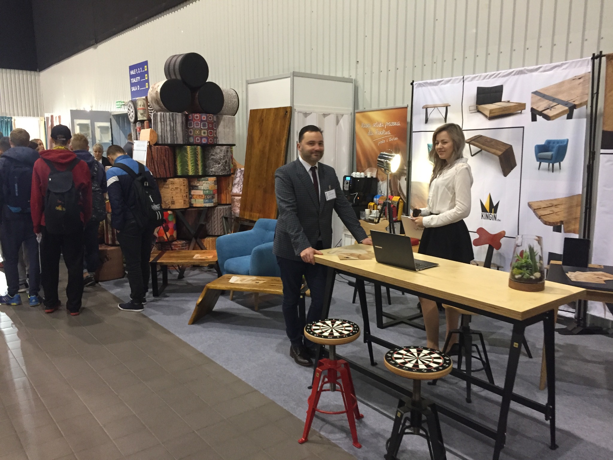 Stoisko targowe z ekspozycją mebli: mężczyzna w garniturze i kobieta przy drewnianym blacie na metalowych nogach, w tle ekspozycja z przykładami mebli i dekoracji, widoczny tłum ludzi.