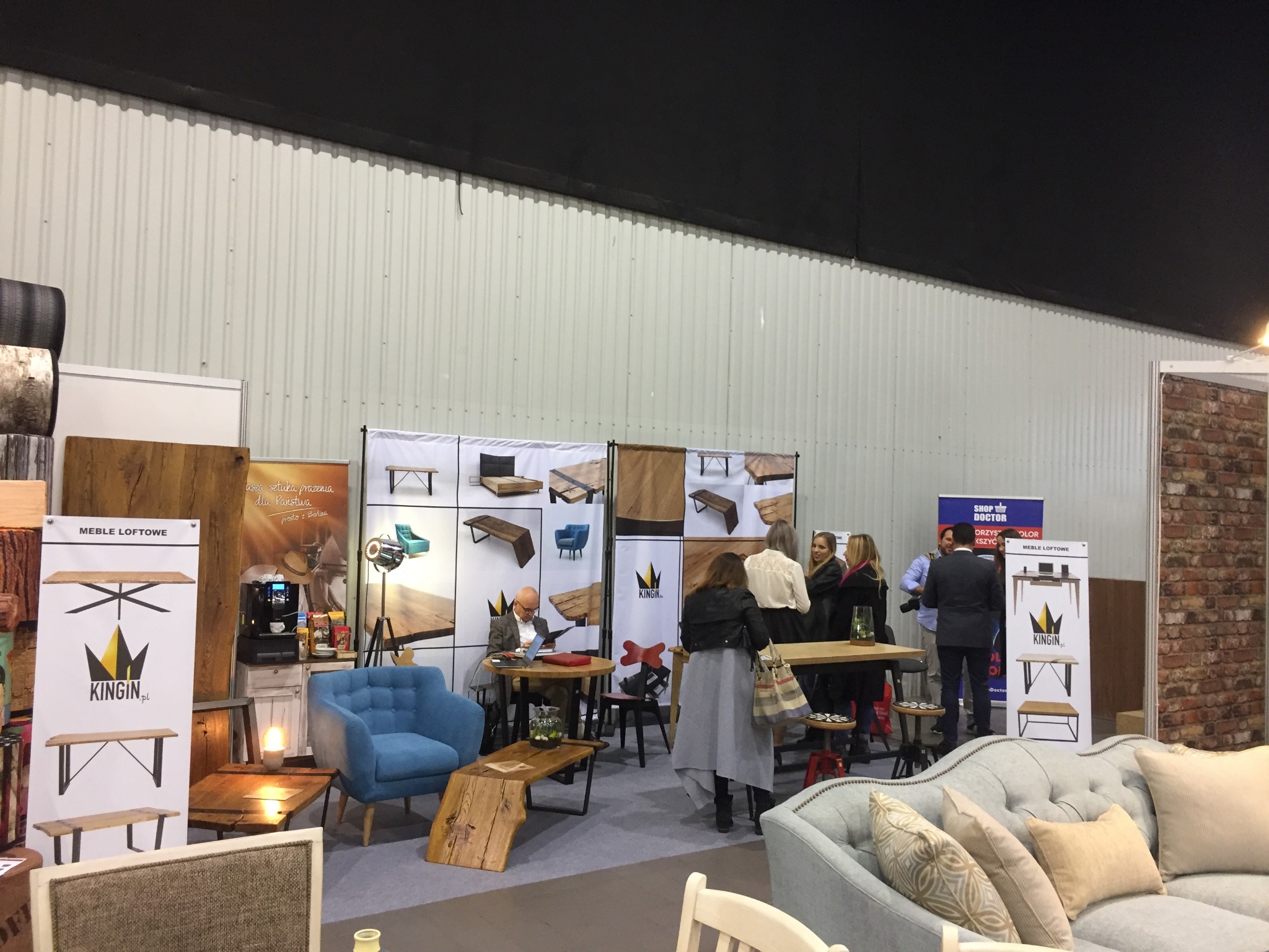 Stoisko targowe z ekspozycją mebli loftowych, w tle osoby oglądające ofertę, na pierwszym planie sofa z poduszkami, widoczny baner reklamowy z logo firmy i prezentacją produktów.
