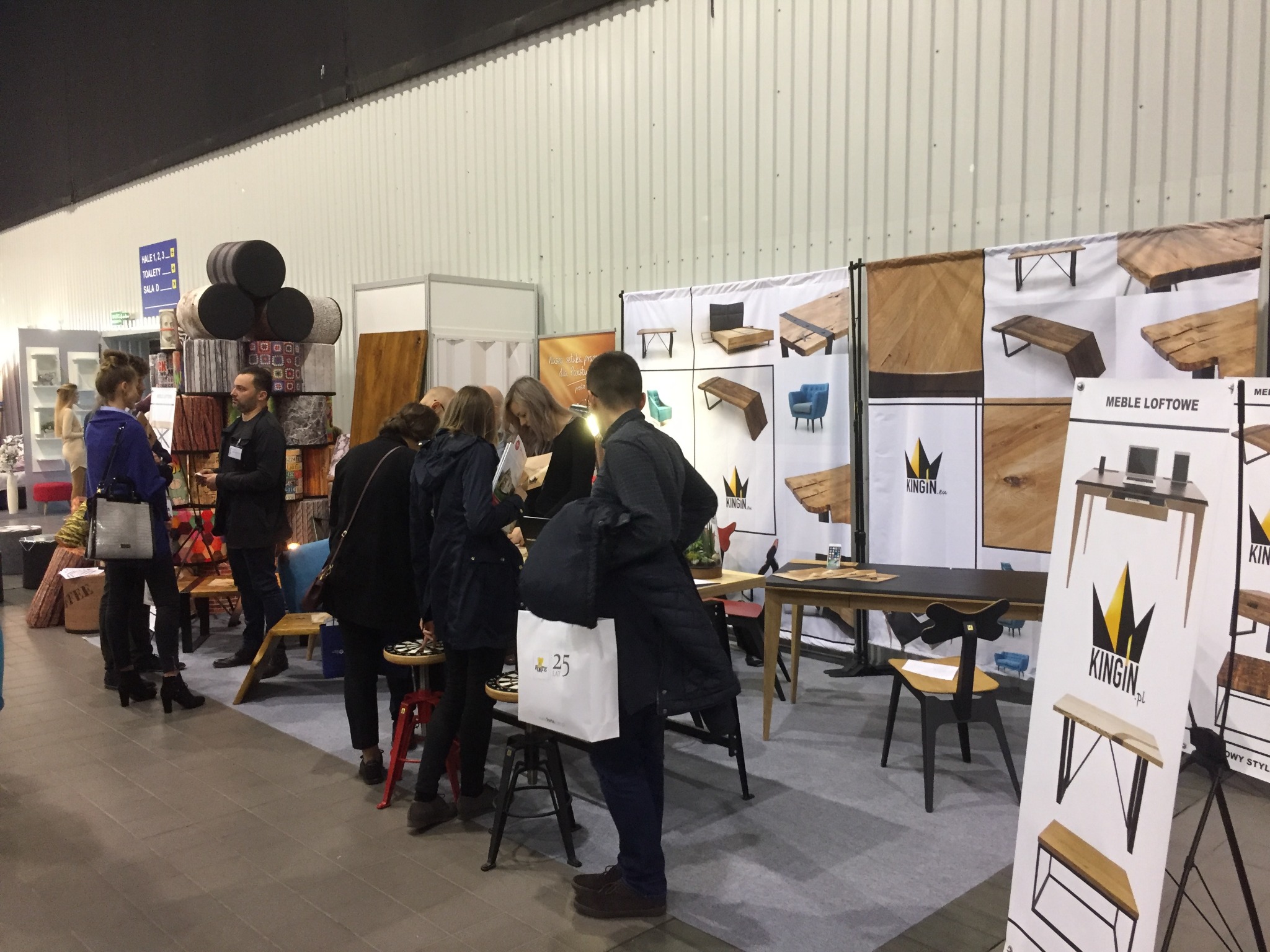 Stoisko targowe z ekspozycją mebli loftowych, prezentujące drewniane stoły, ławy i inne elementy wyposażenia wnętrz. Klienci oglądają katalogi i rozmawiają z przedstawicielami firmy. Na ścianie...