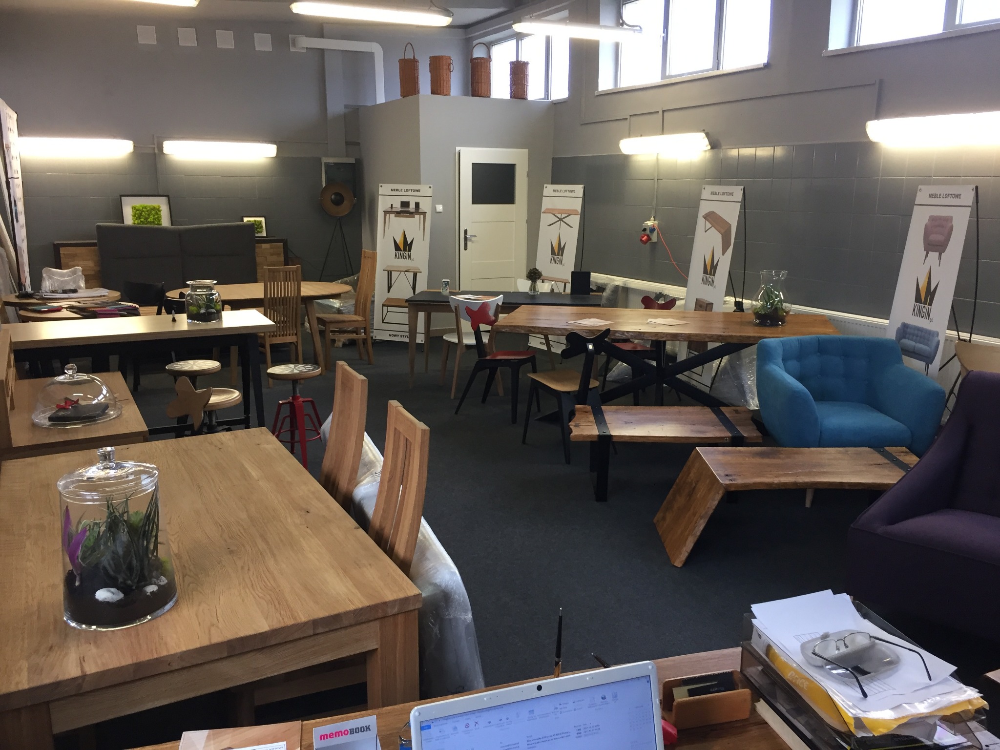 Wnętrze ekspozycji mebli w stylu loftowym, z różnymi stołami, krzesłami i sofami, ustawionymi na ciemnej podłodze; w tle plakaty reklamowe z logo firmy KINGIN i hasłem MEBLE LOFTOWE.