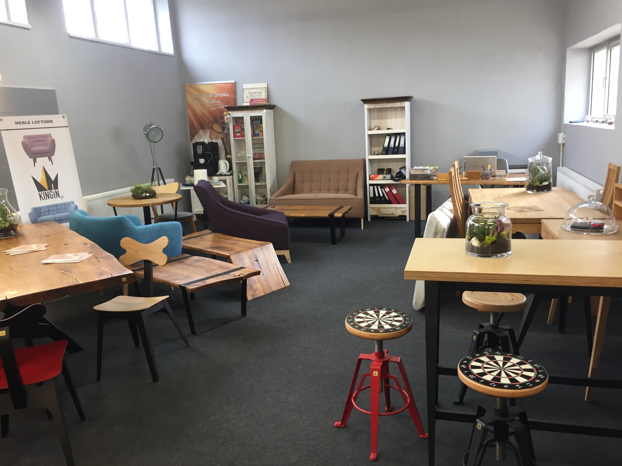 Wnętrze biura lub showroomu z ekspozycją różnych mebli: stoły z drewnianymi blatami, krzesła o różnym designie, sofy, regały, a także dekoracje w postaci terrariów w szklanych naczyniach i stołków...