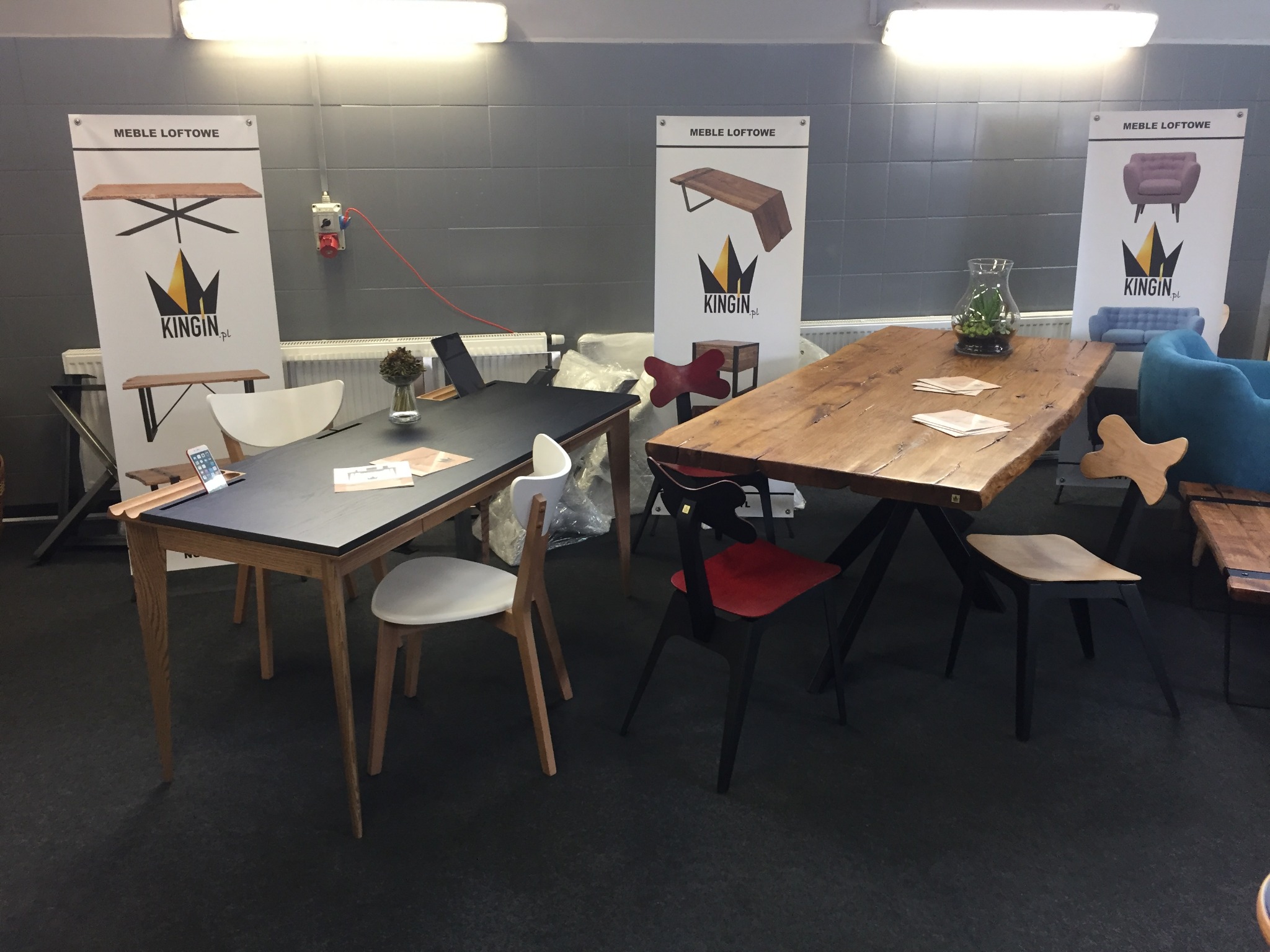 Prezentacja mebli loftowych na targach: stół z czarnym blatem i drewnianymi nogami, stół z blatem z surowego drewna na metalowych nogach, krzesła w różnych stylach, plakaty z logo firmy Kingin...