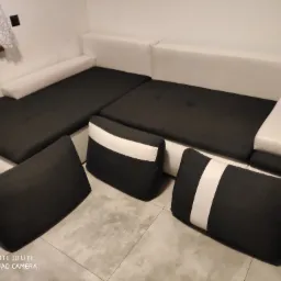 Nowoczesna, narożna kanapa w kolorze białym i grafitowym z trzema poduszkami w pasy, ustawiona na jasnych płytkach.