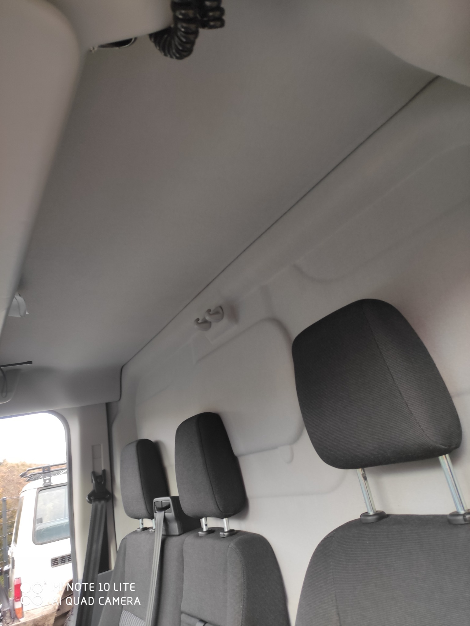 Wnętrze furgonetki z trzema czarnymi zagłówkami widocznymi na tle białej tapicerki, widok z perspektywy pasażera, z fragmentem białego pojazdu widocznym przez otwarte drzwi.