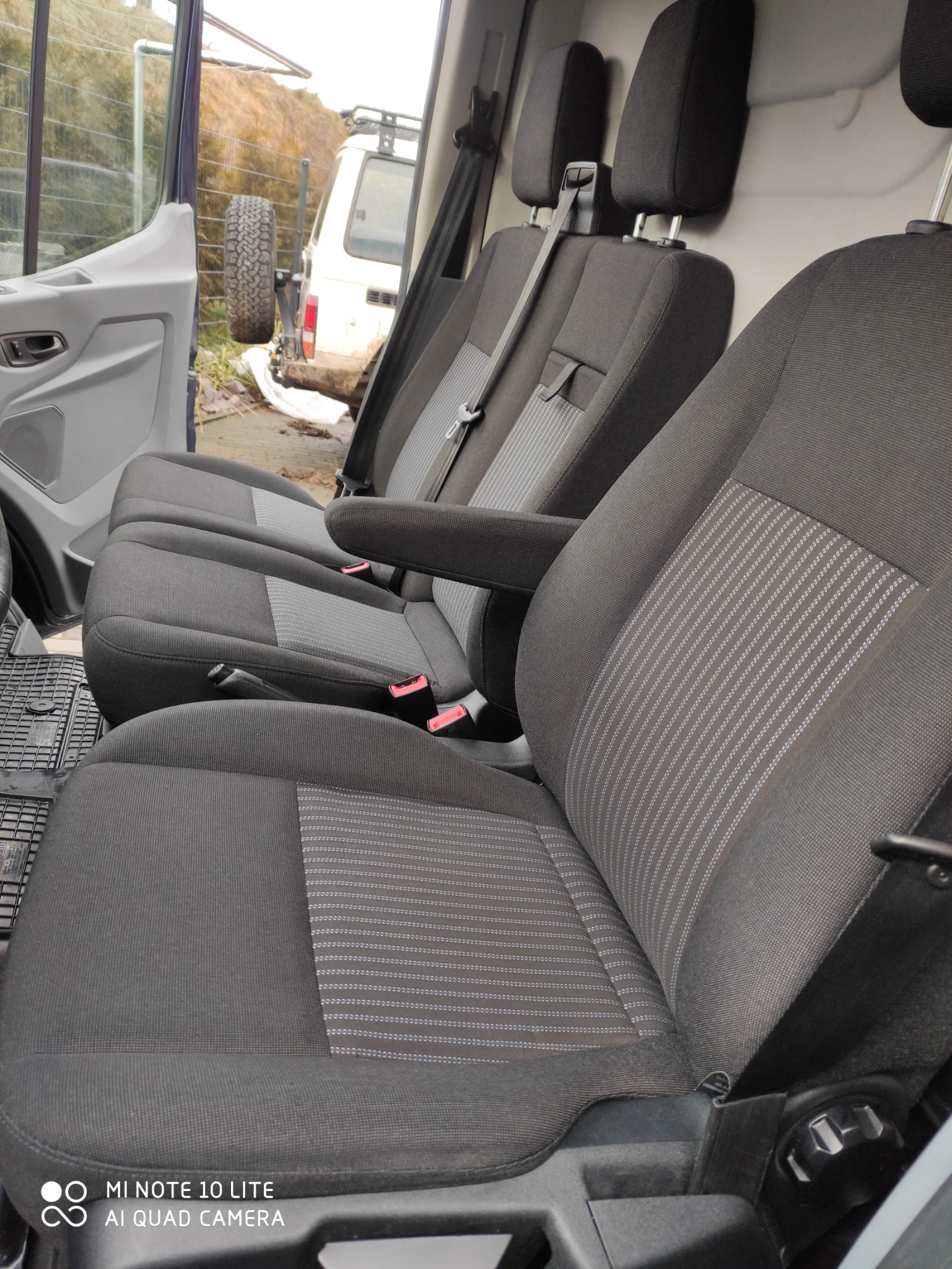 Wnętrze busa z trzema rzędami czarnych foteli z szarymi paskami, widok z otwartych drzwi, na zewnątrz zaparkowany samochód terenowy.