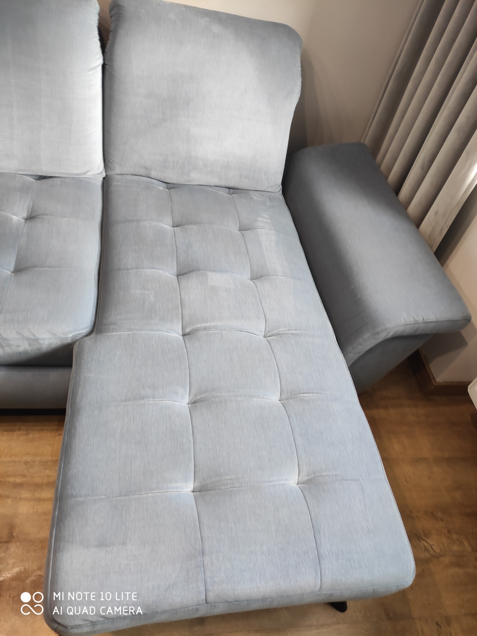 Jasnoszara, rozłożona kanapa z tapicerką w kwadraty, widoczne plamy po czyszczeniu, częściowo zasłonięte zasłony w tle.