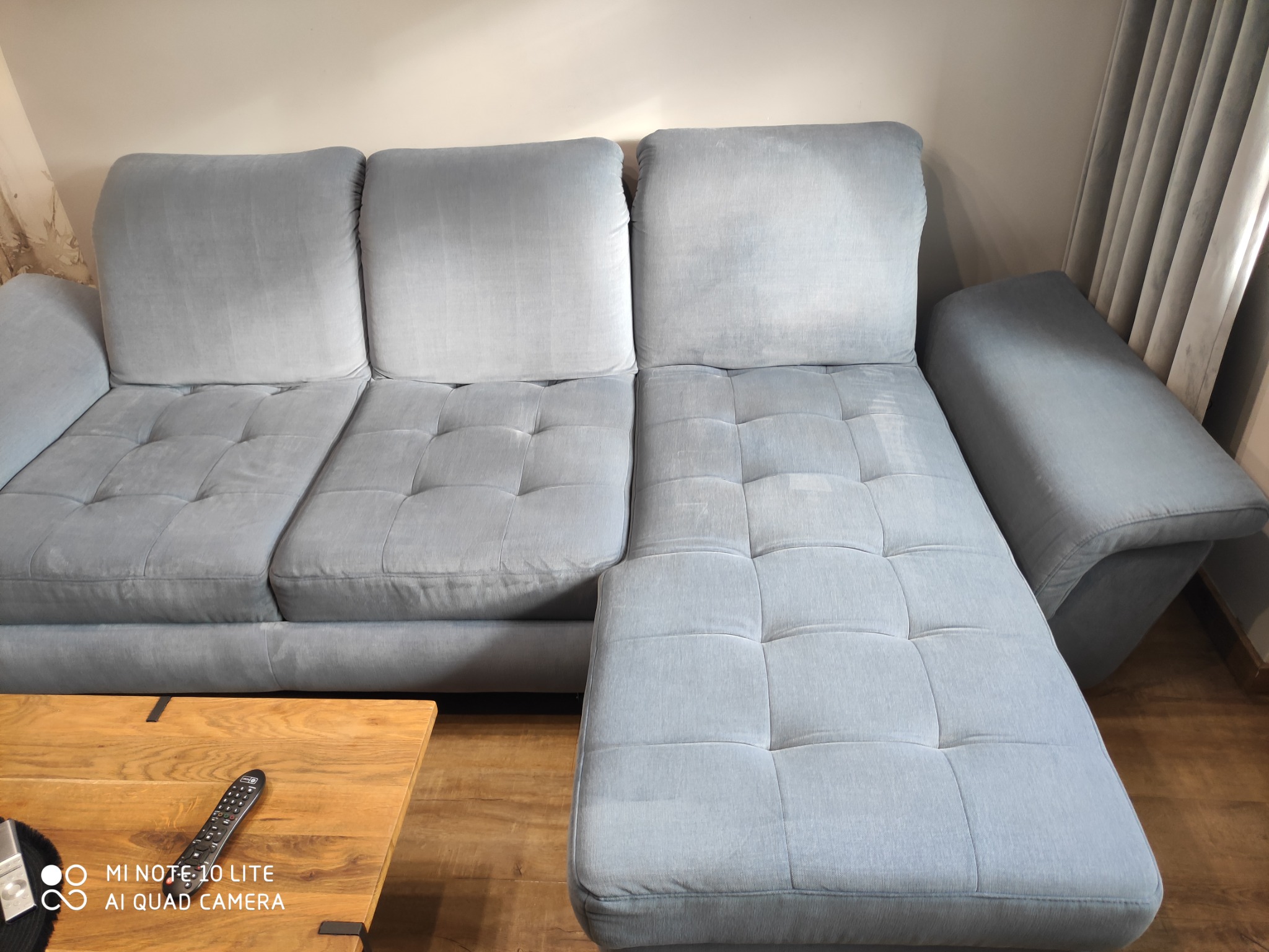 Jasnoszara, modułowa kanapa z pikowanymi siedziskami i oparciami, widoczna z niskiej perspektywy, na drewnianej podłodze, z pilotem leżącym na drewnianym stoliku kawowym.