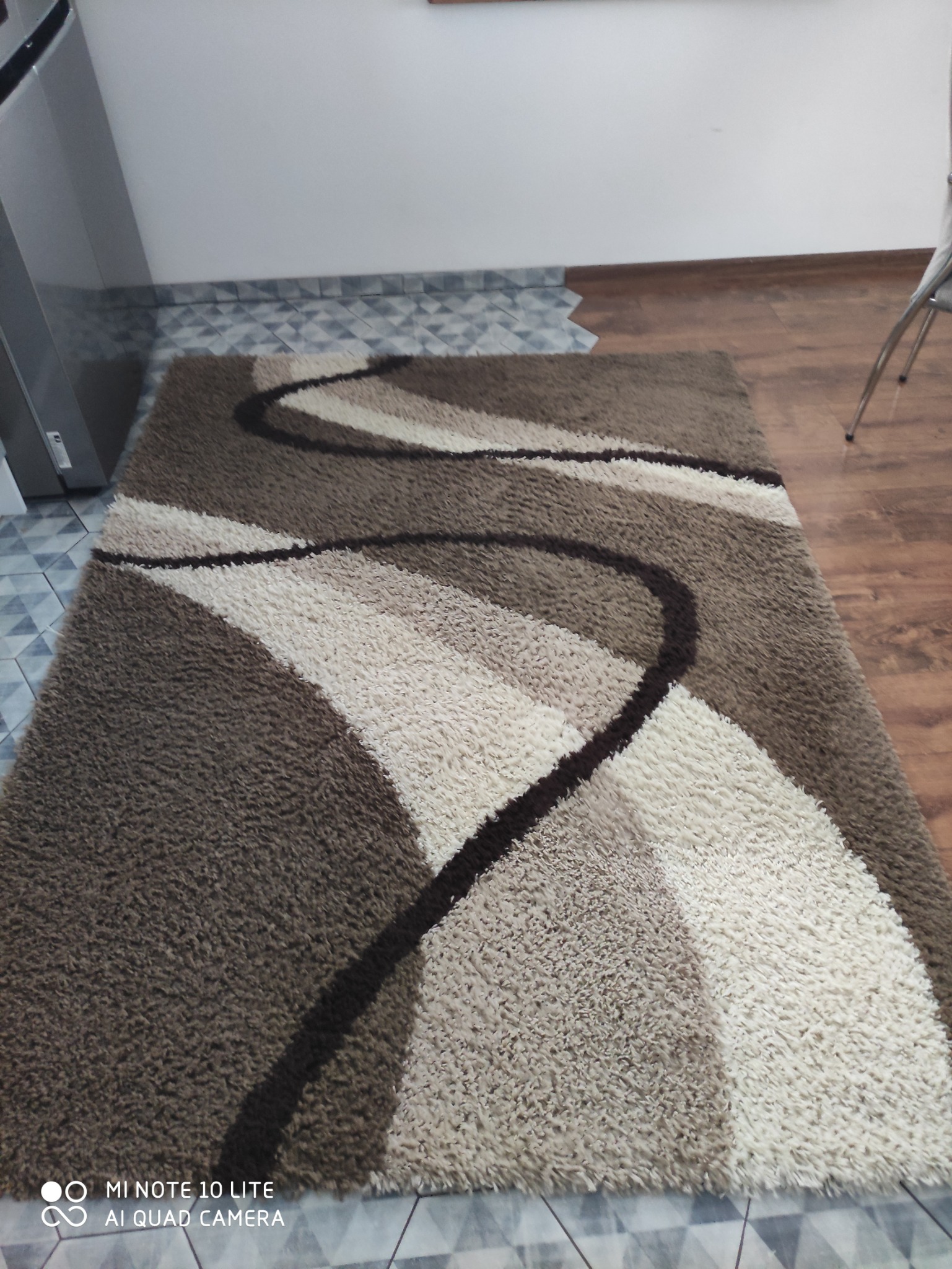 Wzorzysty dywan shaggy w odcieniach brązu i beżu z ciemnobrązowym motywem falistym, leżący na podłodze z płytek imitujących mozaikę i drewnianej podłodze.