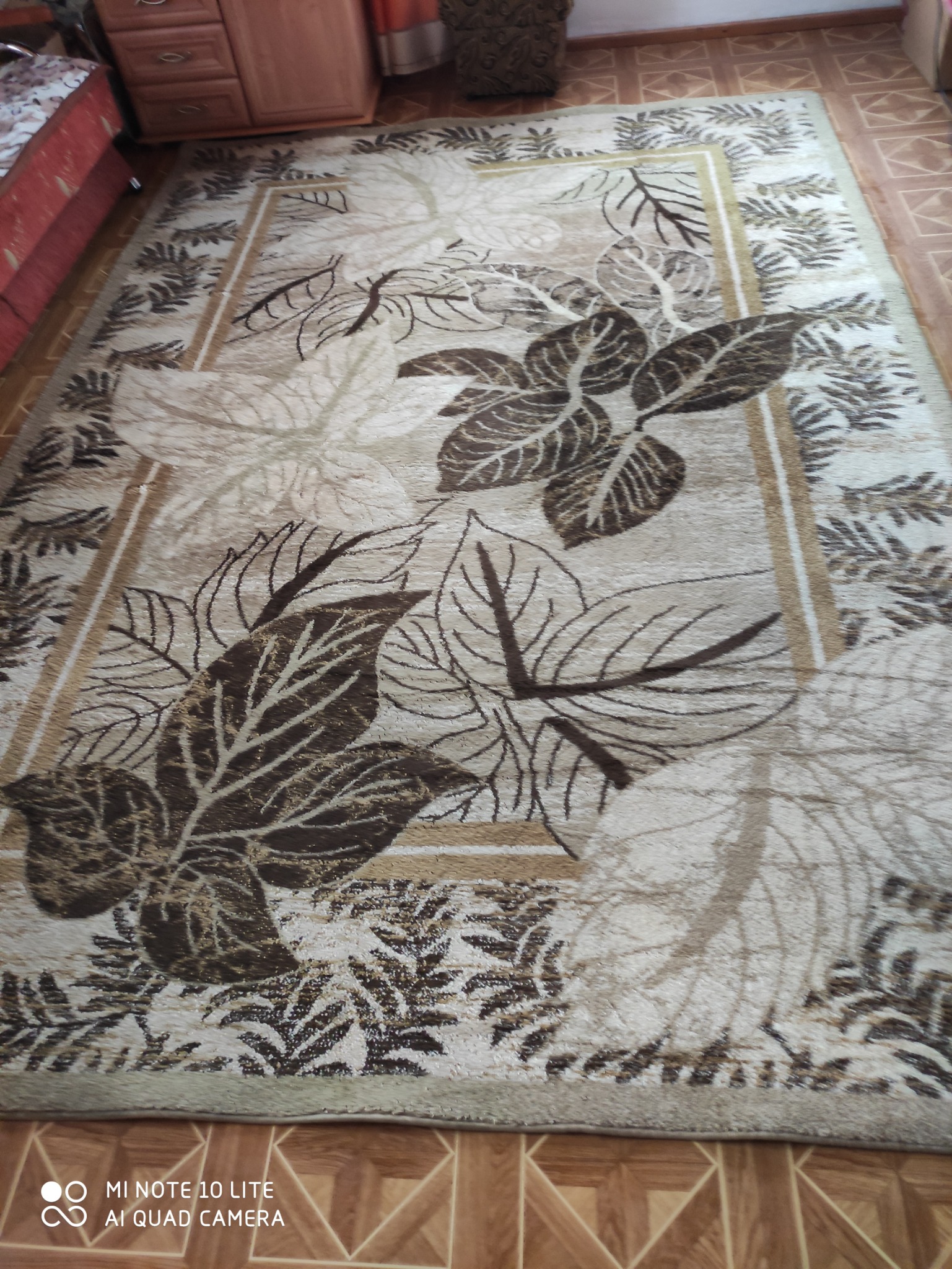 Prostokątny dywan z motywem liści w odcieniach brązu i beżu, leżący na podłodze z brązowych płytek.