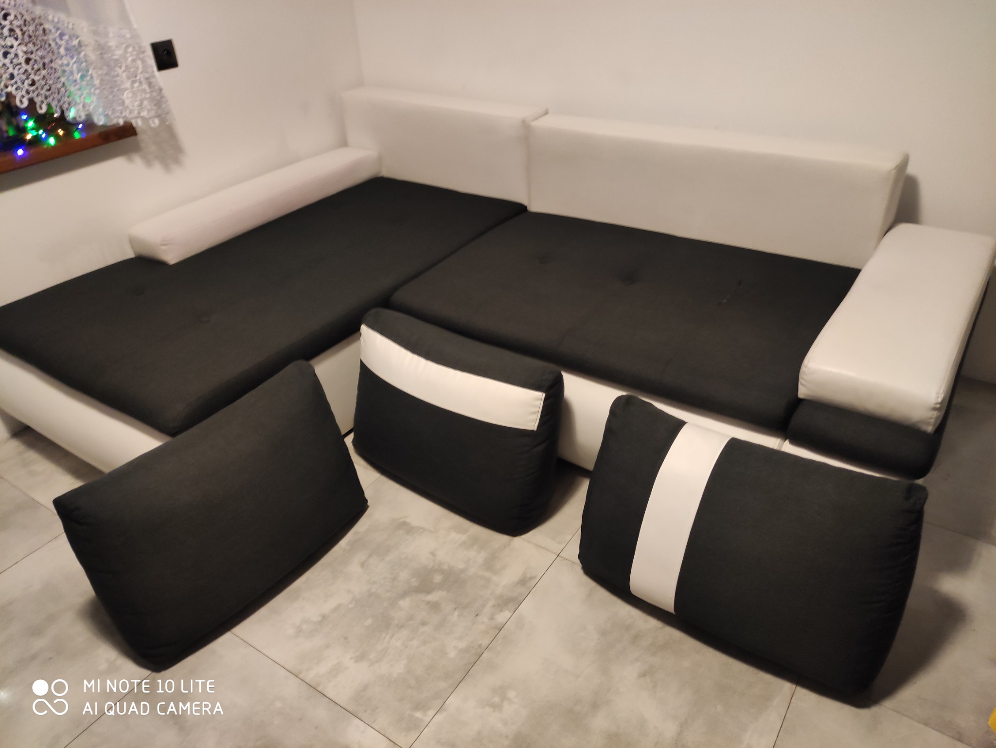 Nowoczesna, narożna kanapa w kolorze białym i grafitowym z trzema poduszkami w pasy, ustawiona na jasnych płytkach.