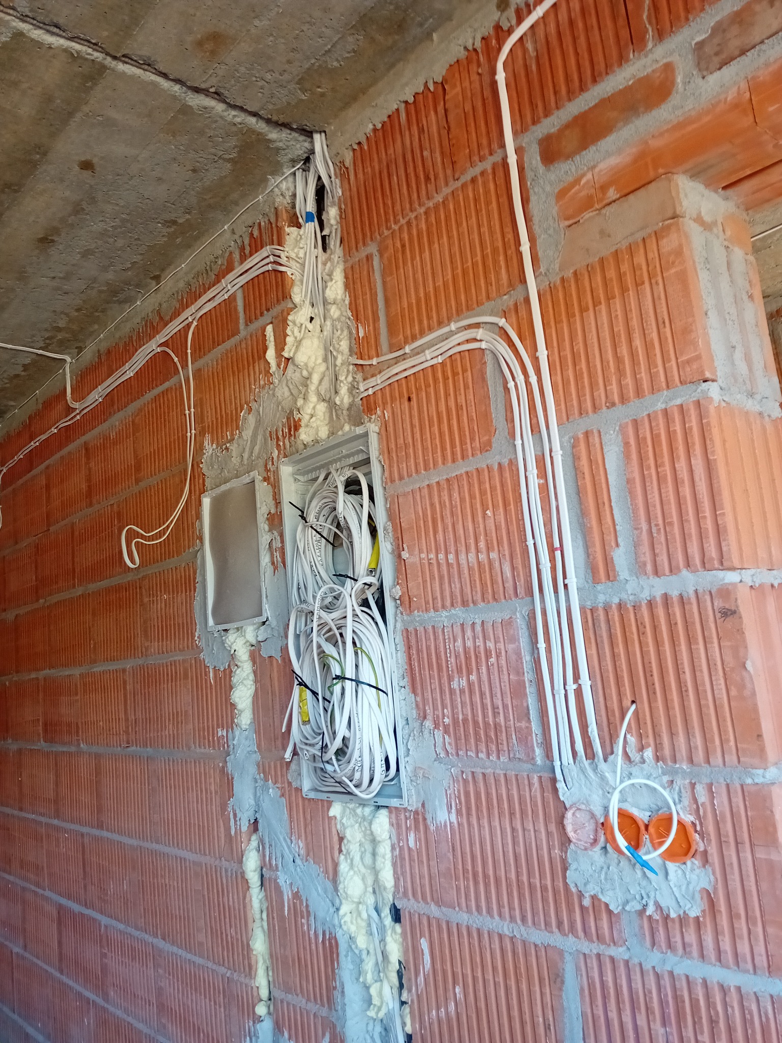 Rozprowadzenie przewodów elektrycznych w ścianie z pustaków ceramicznych, zabezpieczone pianką montażową, widoczna puszka instalacyjna z licznymi kablami.