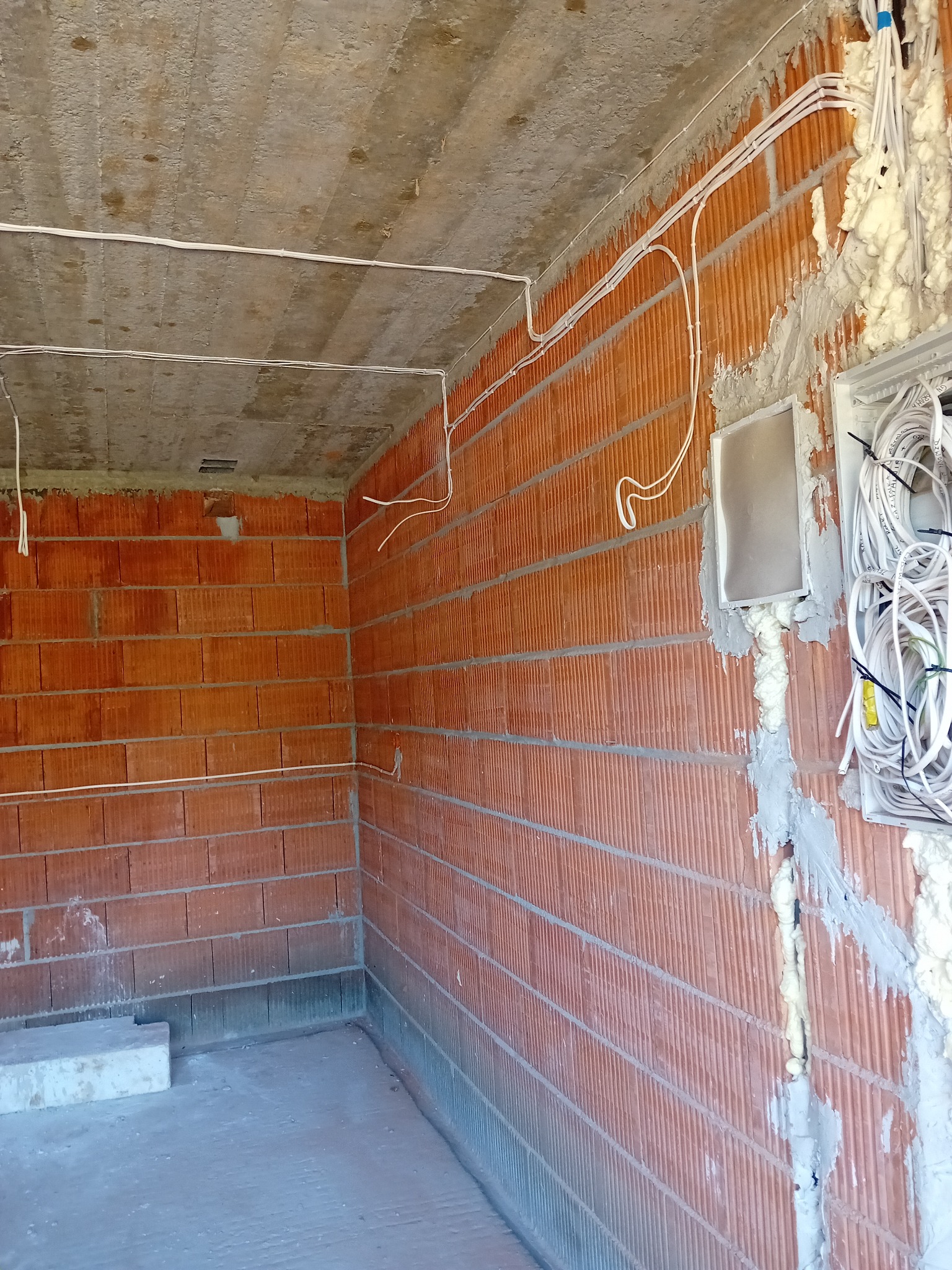 Surowy stan instalacji elektrycznej w budynku z czerwonej cegły, widoczne kable prowadzone po ścianach i pod sufitem, puszki elektryczne osadzone w pianie montażowej.