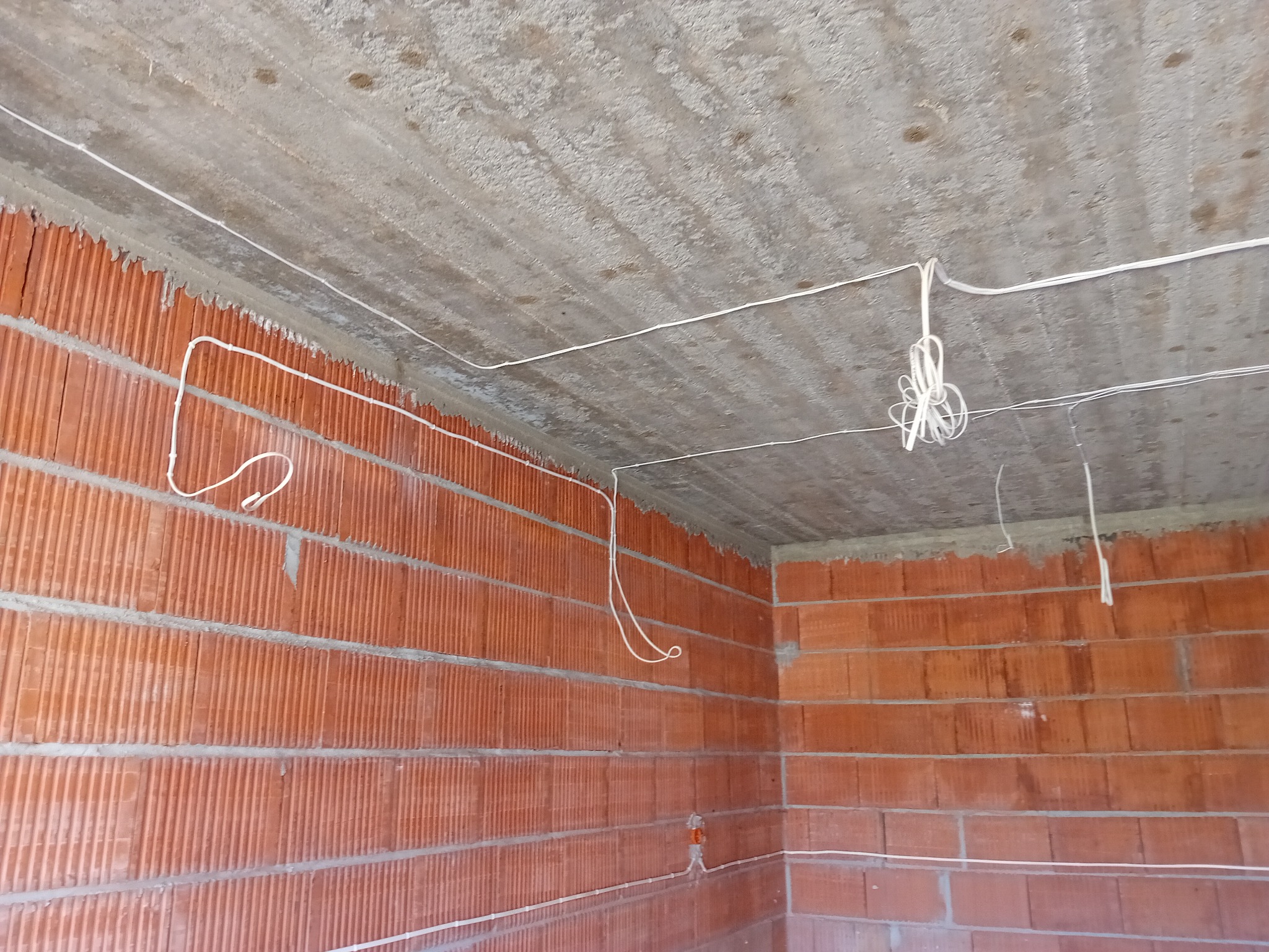 Surowa ściana z czerwonej cegły i betonowy sufit z poprowadzonymi przewodami elektrycznymi, widoczne pętle kabli zwisające z sufitu.