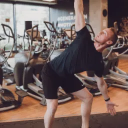 Mężczyzna w czarnym stroju sportowym wykonuje ćwiczenie z kettlebellem w siłowni, z maszynami eliptycznymi w tle.