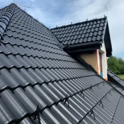 Błyszczący, czarny dach z dachówki betonowej po malowaniu, z widocznymi zabezpieczeniami przeciwśniegowymi i anteną na szczycie, na tle błękitnego nieba z chmurami.