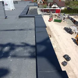 Widok z góry na płaski dach pokryty papą, z widocznymi obróbkami blacharskimi, workiem izolacji IZObud i placem budowy w tle.