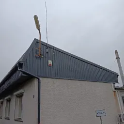 Szary dach z blachy trapezowej na budynku z elewacją w jasnym kolorze, widoczna rynna, lampa uliczna i antena na dachu, tabliczka z napisem 'ZAOPATRZENIE'.