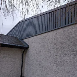 Fragment budynku z szarą blachą trapezową na górnej części ściany, widoczne również fragmenty dachu pokrytego papą i rynny, na pierwszym planie gałęzie drzewa.