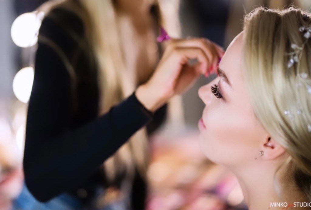 Profil kobiety podczas aplikacji pędzlem kosmetyku na brwi, widoczne sztuczne rzęsy i delikatne upięcie włosów z ozdobami