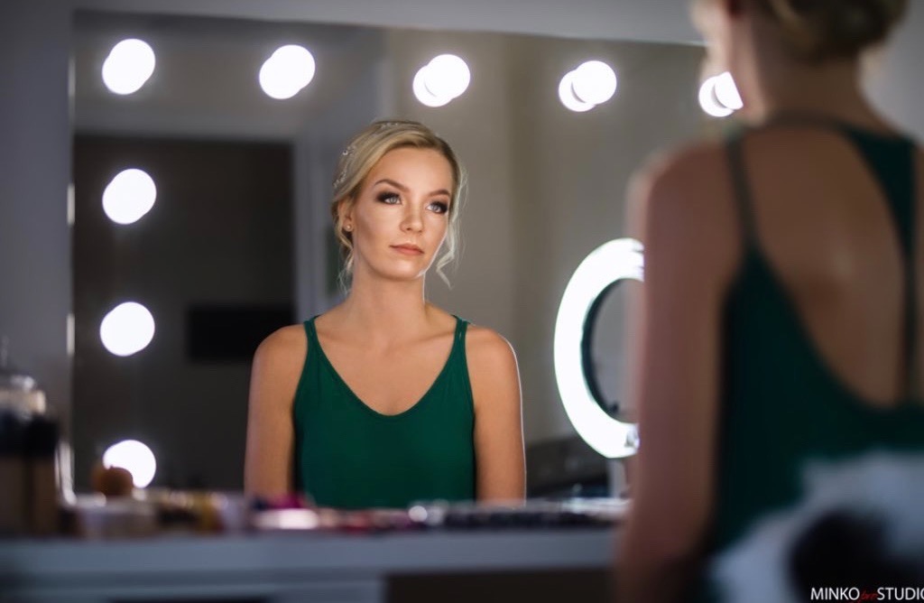 Portret kobiety z delikatnym makijażem, widoczna w lustrze z oświetleniem punktowym, w tle ring lampa