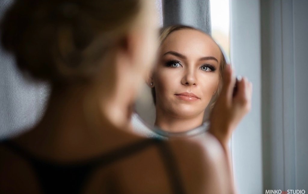 Portret kobiety po zabiegu upiększającym, odbicie w okrągłym lustrze, widoczne starannie wykonane cieniowanie powiek i błyszczące usta, jasna karnacja.