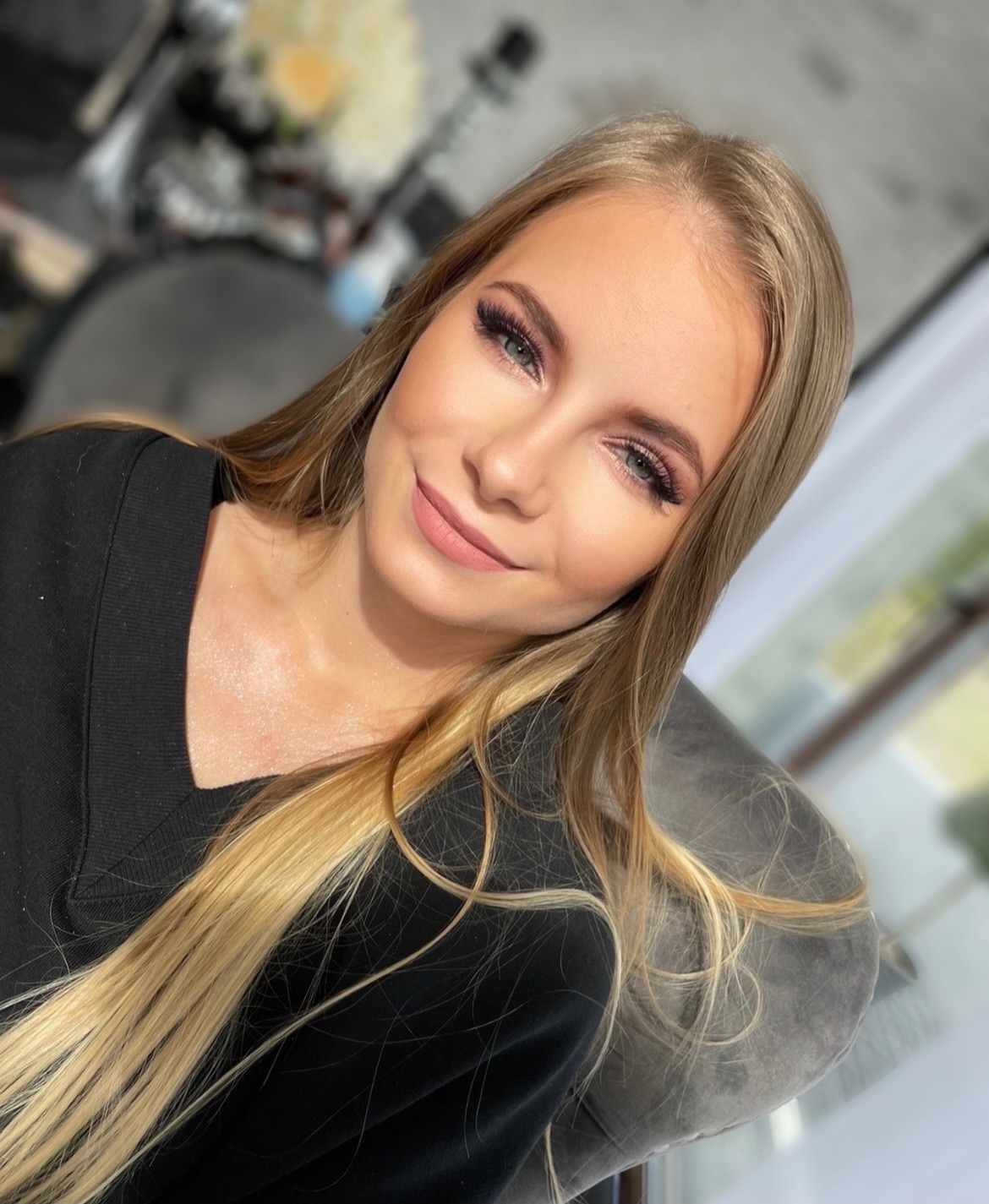 Portret uśmiechniętej blondynki z delikatnym, różowym makijażem oczu i ust, sztucznymi rzęsami, widoczne refleksy na włosach i dekolcie.