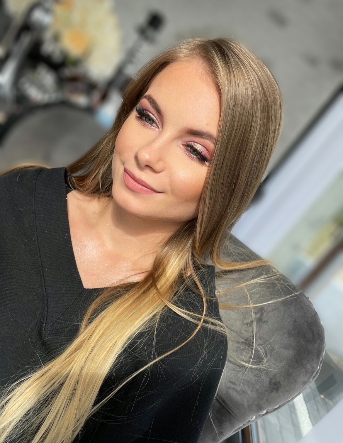 Portret uśmiechniętej blondynki z delikatnym makijażem w odcieniach różu i beżu, z widocznym rozświetlaczem na policzkach, na tle rozmytego wnętrza salonu.