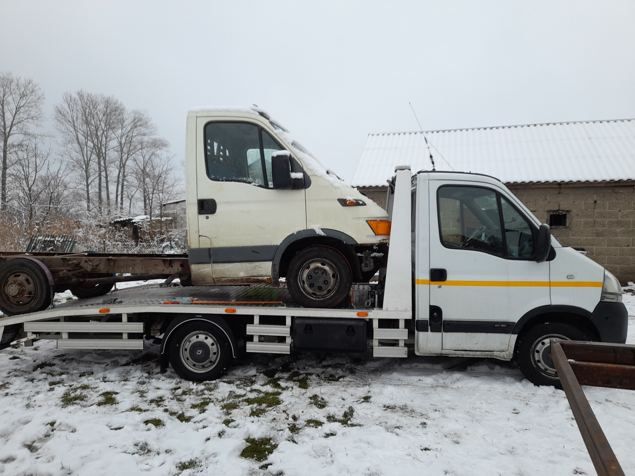 Uszkodzony biały van bez karoserii przewożony na platformie transportowej w zimowej scenerii, z widocznymi śladami śniegu na pojeździe i ziemi.