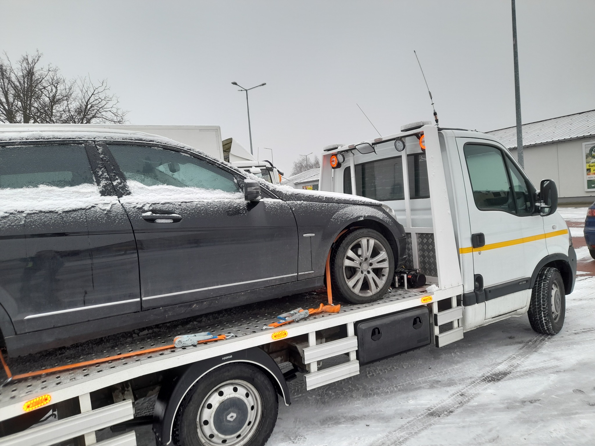Czarny samochód osobowy, pokryty śniegiem, transportowany na białej platformie pojazdu pomocy drogowej z żółtą linią, w zimowych warunkach atmosferycznych.