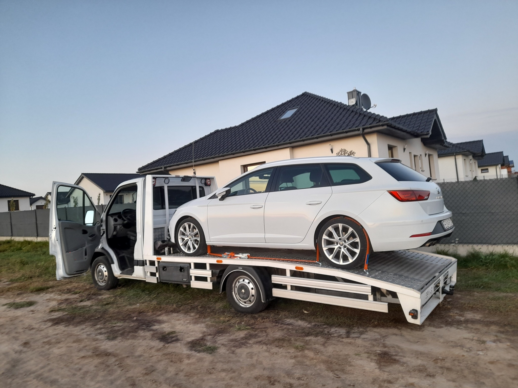 Biała platforma transportowa z otwartymi drzwiami kabiny, na której zabezpieczony jest biały samochód osobowy typu kombi, na tle osiedla domów jednorodzinnych.