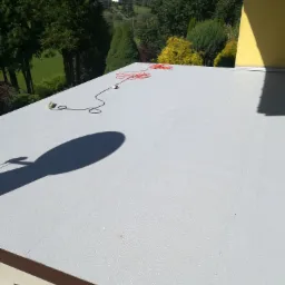 Szary, płaski dach pokryty papą termozgrzewalną z widocznym kablem elektrycznym i cieniem narzędzia, w tle zieleń i budynek.