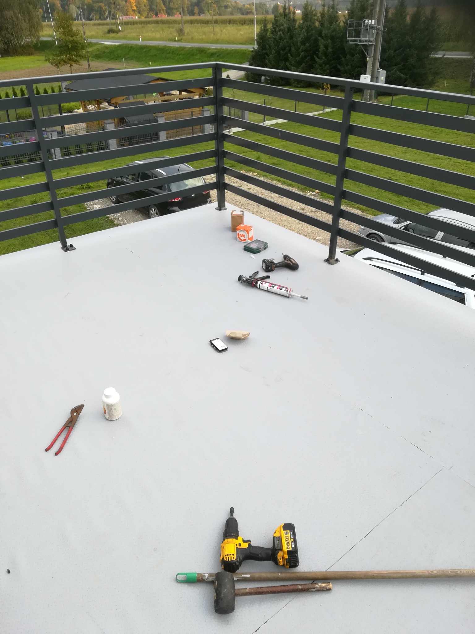 Widok z góry na płaski dach z narzędziami i materiałami budowlanymi: wiertarka, pistolet do uszczelniania, szczypce, puszka, telefon, paczka, widoczne barierki i otoczenie budynku.