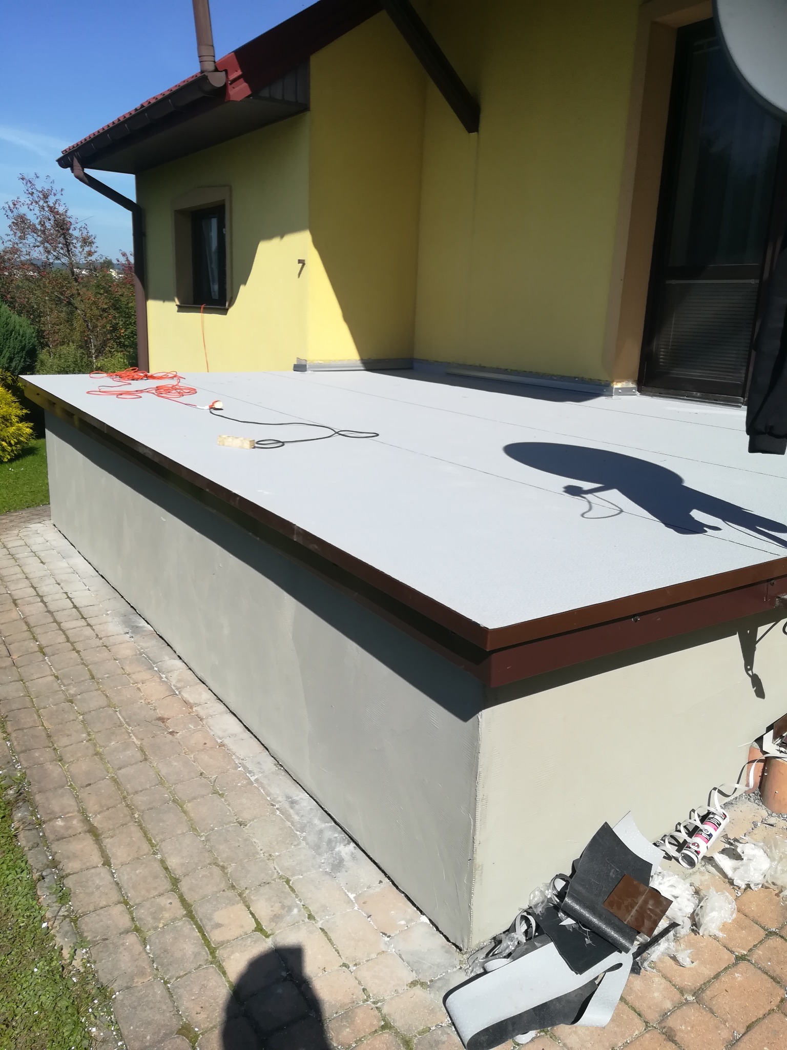 Wykończony płaski dach z szarą membraną i brązowymi obrzeżami, widoczne narzędzia i resztki materiałów po montażu, na tle żółtej ściany budynku z numerem 7 i fragmentem ogrodu.