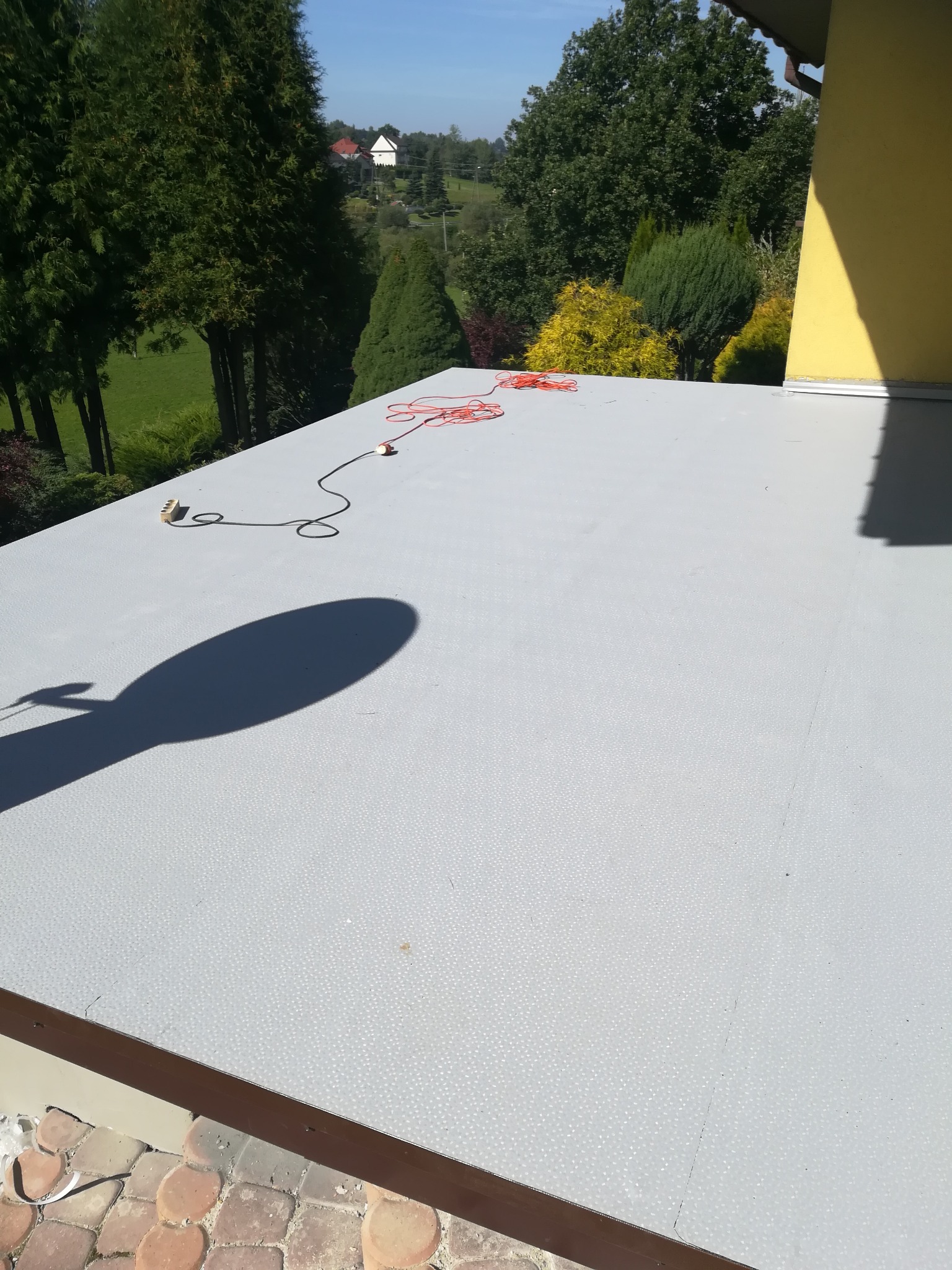 Szary, płaski dach pokryty papą termozgrzewalną z widocznym kablem elektrycznym i cieniem narzędzia, w tle zieleń i budynek.