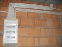 Rozprowadzenie przewodów elektrycznych w białych peszlach, prowadzących do skrzynki rozdzielczej zamontowanej na ścianie z czerwonej cegły dziurawki w surowym stanie.