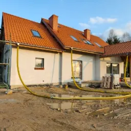 Dom jednorodzinny w trakcie prac elewacyjnych z widocznymi rusztowaniami, żółtymi wężami doprowadzającymi materiał i częściowo wykończoną elewacją, teren wokół domu podmokły.