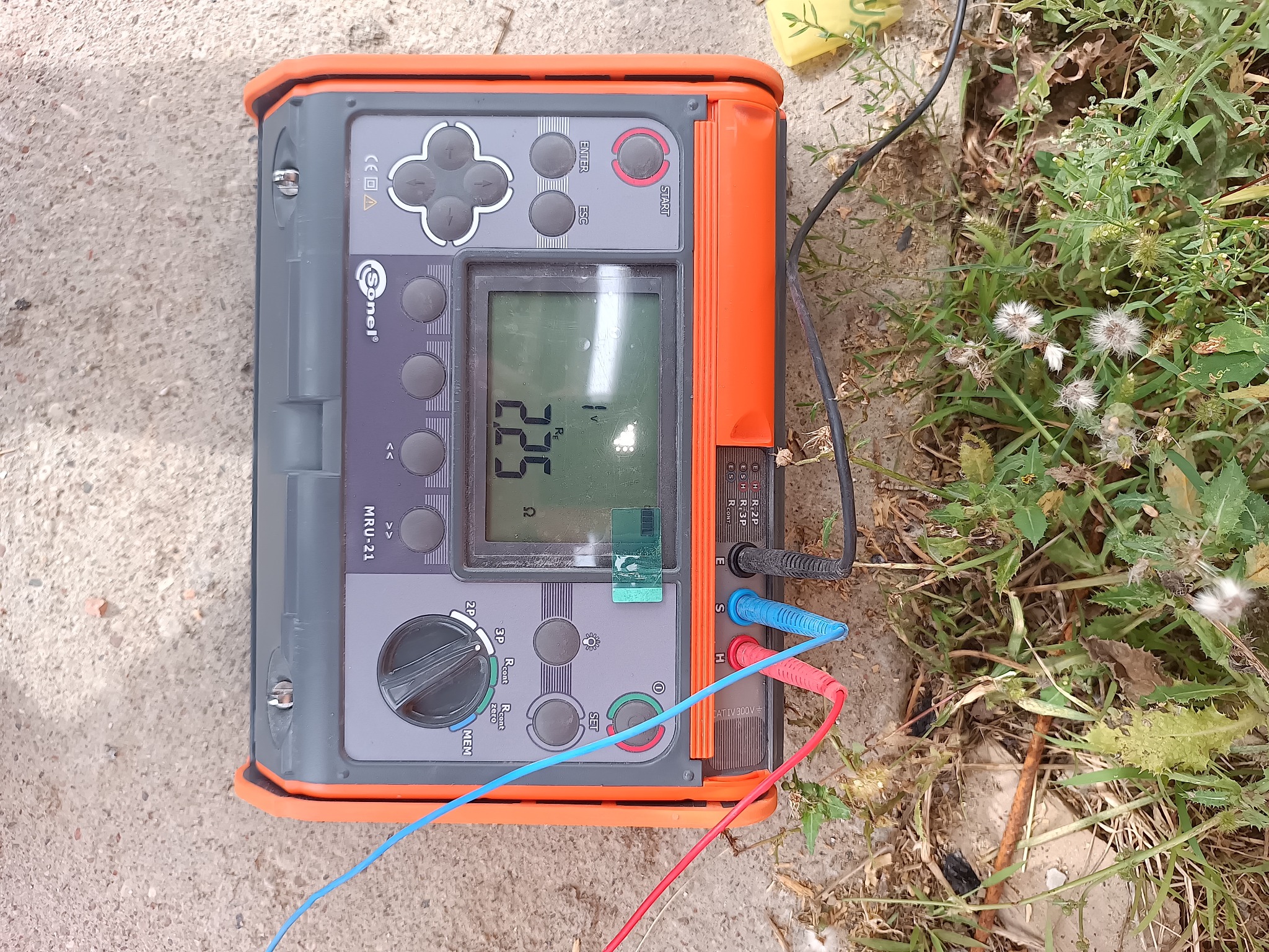 Miernik Sonel MRU-21 z podłączonymi przewodami pomiarowymi, leżący na ziemi z trawą. Urządzenie wskazuje wartość 225.