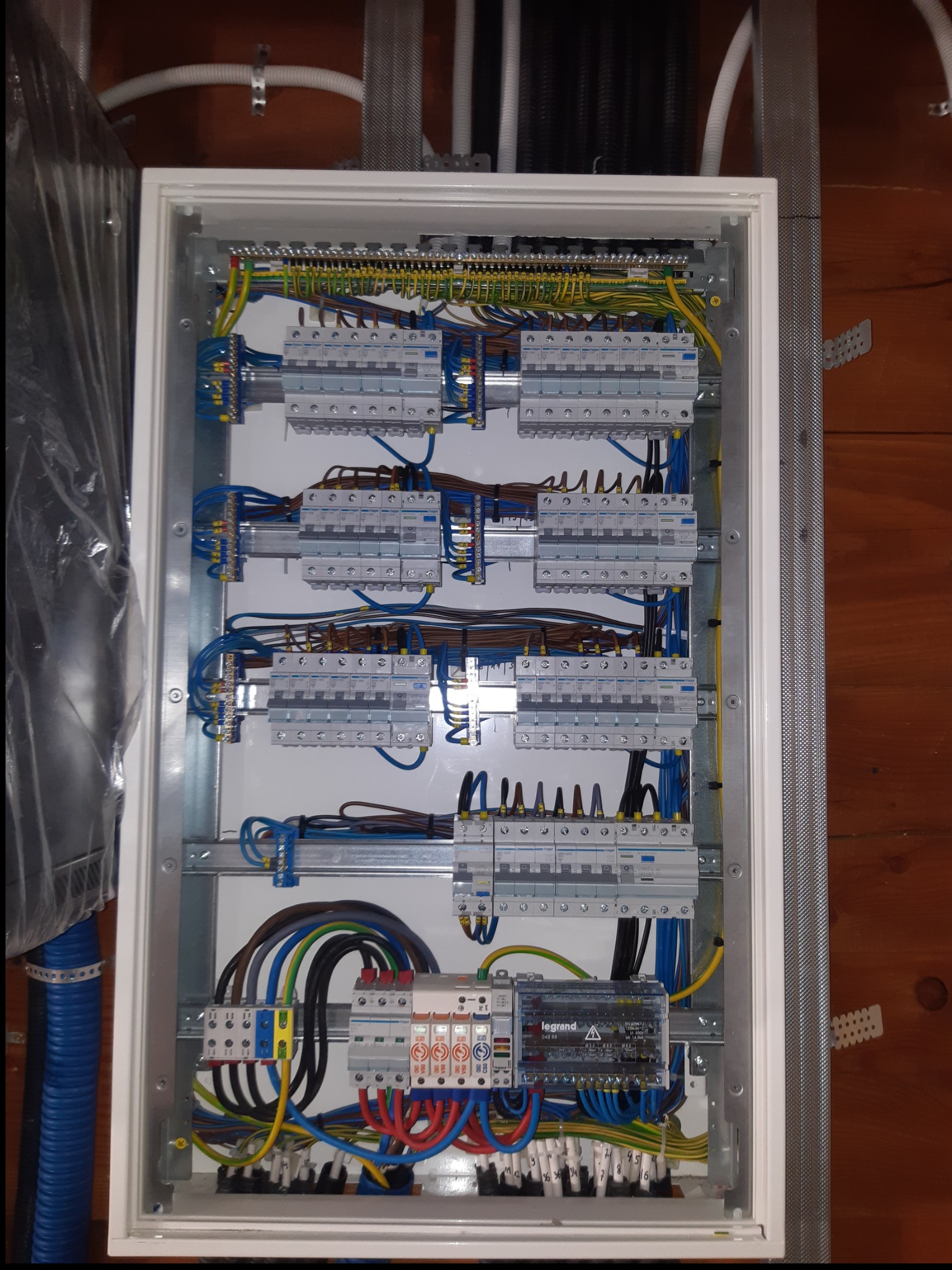 Otwarta skrzynka rozdzielcza z bezpiecznikami i okablowaniem, widoczne brązowe panele ścienne w tle, niebieska rura karbowana po lewej stronie.