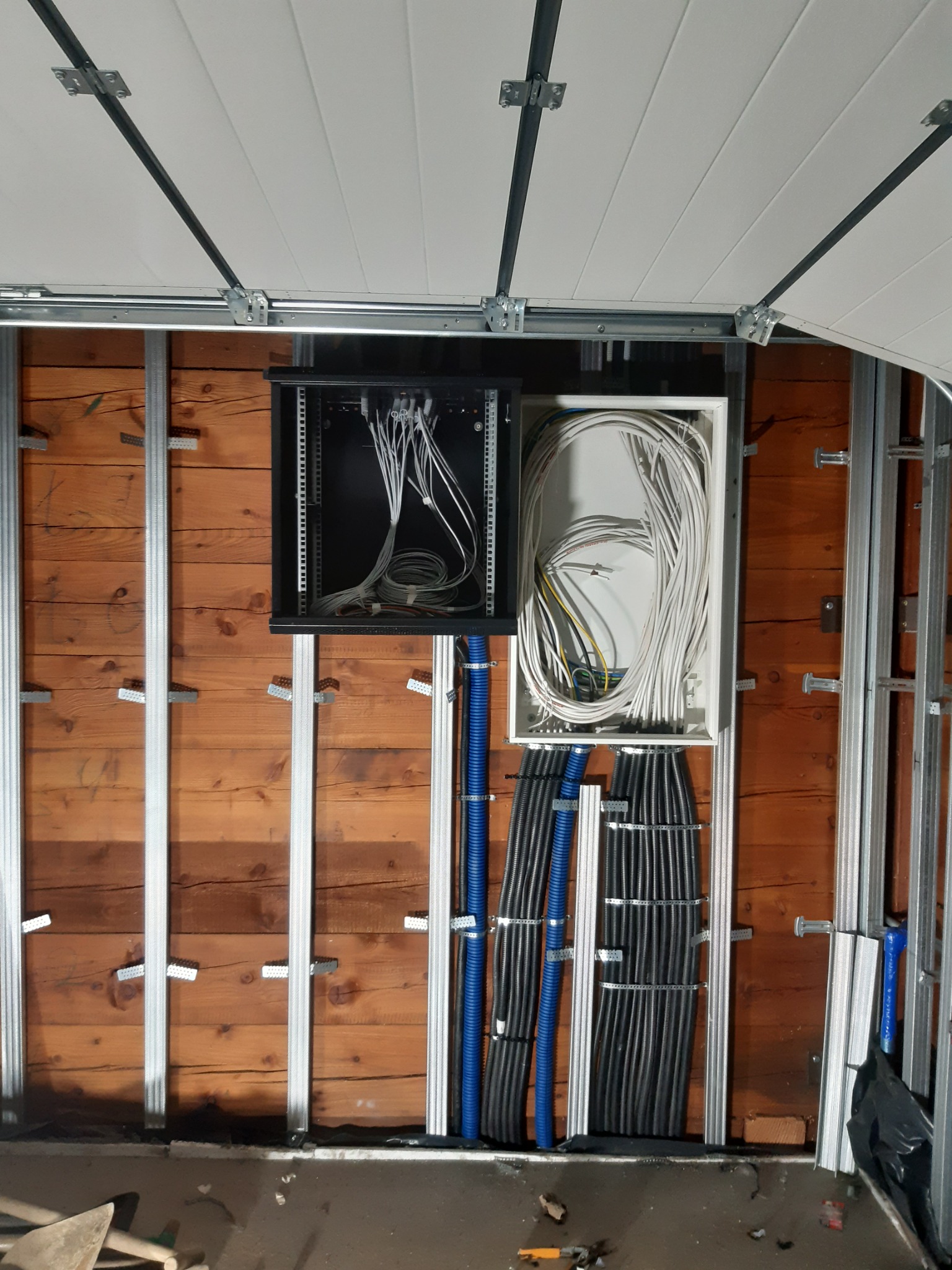 Rozdzielnia elektryczna z przewodami w peszlach, zamontowana na drewnianej ścianie, widoczne metalowe profile konstrukcyjne i elementy mocujące.