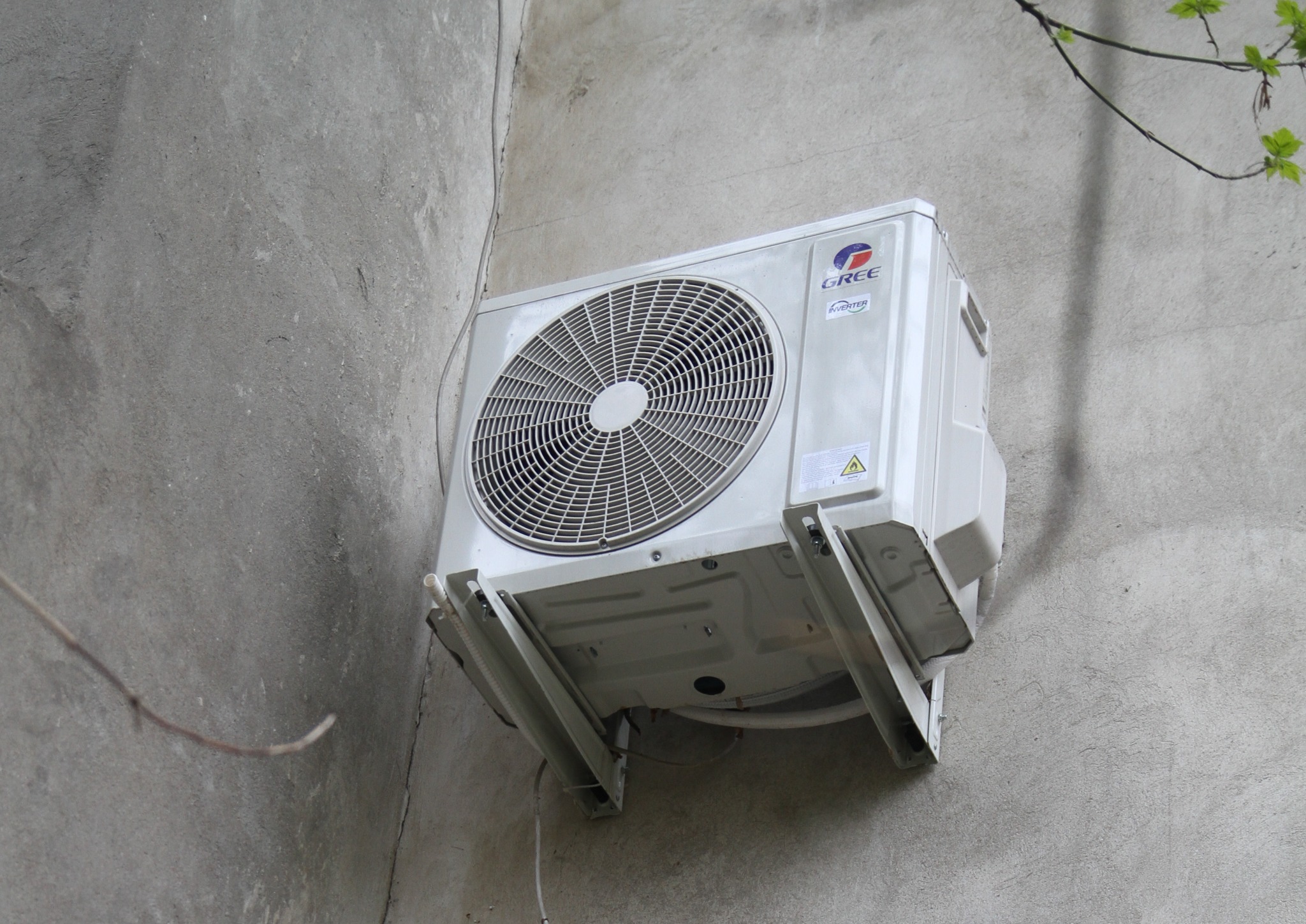 Biała jednostka zewnętrzna klimatyzacji marki Gree, zamontowana na szarym, chropowatym murze z widocznymi przewodami i fragmentem gałęzi drzewa w tle.
