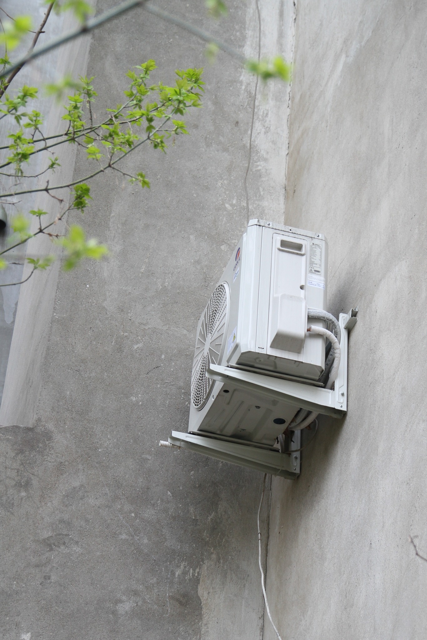 Zewnętrzna jednostka klimatyzacji zamontowana na wsporniku na szarej ścianie budynku, widoczne kable i rurki, z lewej strony fragment drzewa z młodymi liśćmi.