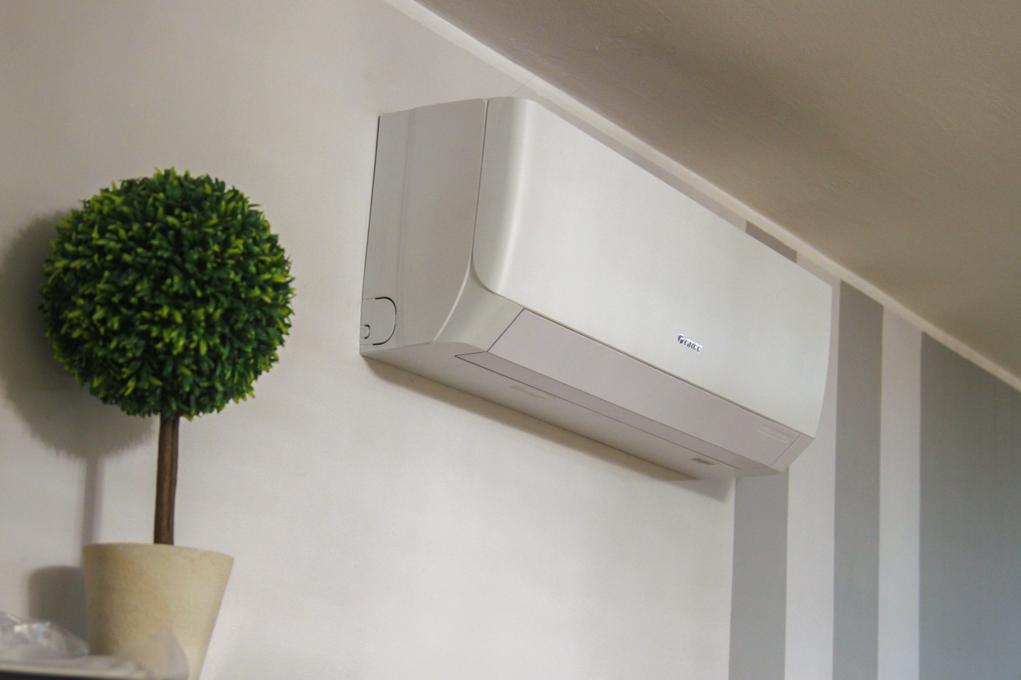 Biała klimatyzacja marki Gree zamontowana na ścianie z dekoracyjnymi szarymi pasami, obok sztuczna roślina w doniczce.