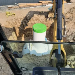 Widok z kabiny koparki na wykop z wstawionym zbiornikiem z zieloną pokrywą, w tle widoczna ziemia i materiały budowlane.