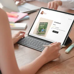 Osoba przegląda ofertę e-booka kulinarnego na tablecie z klawiaturą, używając rysika; na drewnianym biurku leżą zakreślacze i doniczka z zieloną rośliną.