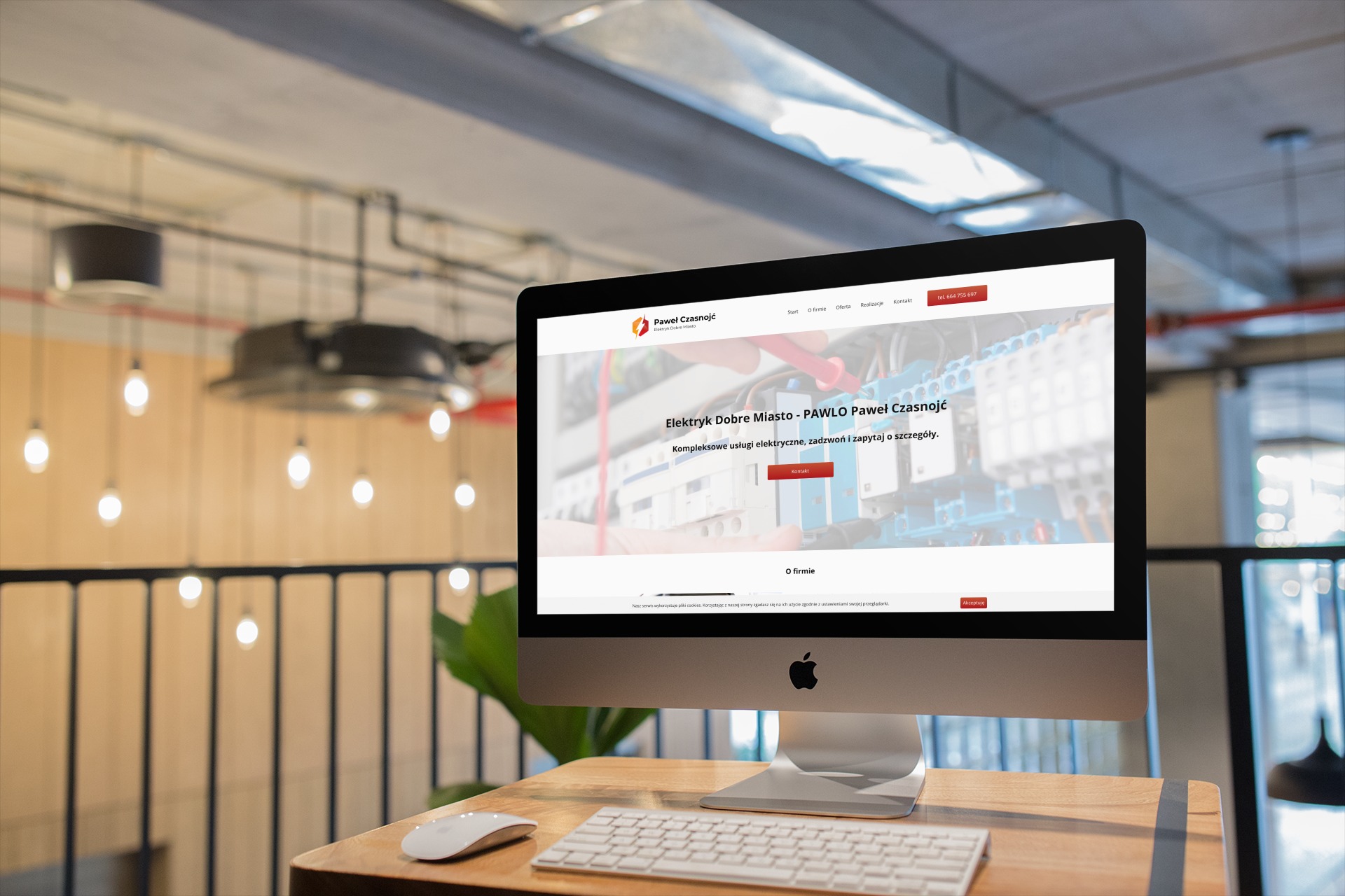 Ekran komputera iMac wyświetlający stronę internetową firmy elektrycznej 'Paweł Czasnojć', z widocznym logo, menu nawigacyjnym i hasłem reklamowym na tle wnętrza biura z designerskim oświetleniem.