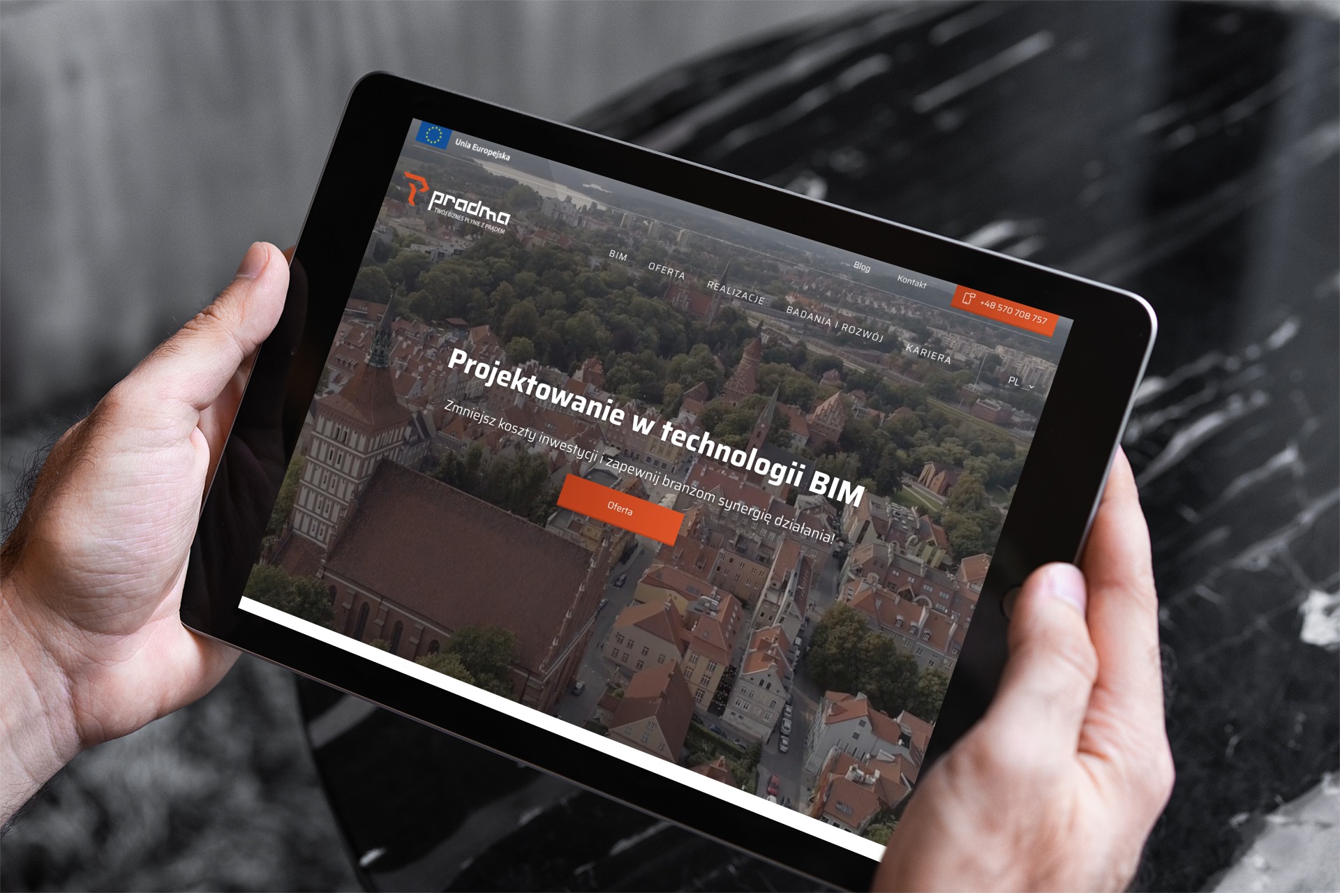 Mężczyzna trzyma tablet prezentujący stronę internetową firmy Pradma z ofertą projektowania w technologii BIM, na tle widok na miasto.